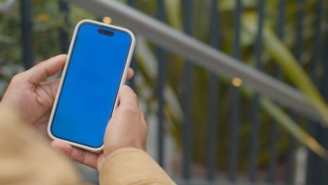 Close-Up-Of-Muslim-Man-Sitting-Outdoors-On-City-Street-Looking-At-Blue-Screen-Mobile-Phone-2
